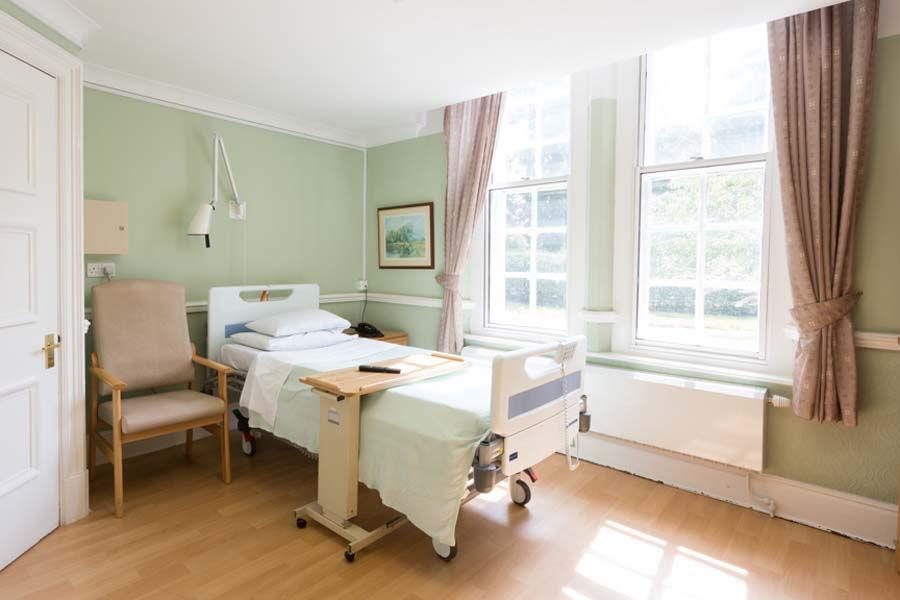A single hospital bed with an overbed table in a pale green room. Sunlight comes through tall windows, and an empty armchair stands beside the bed.