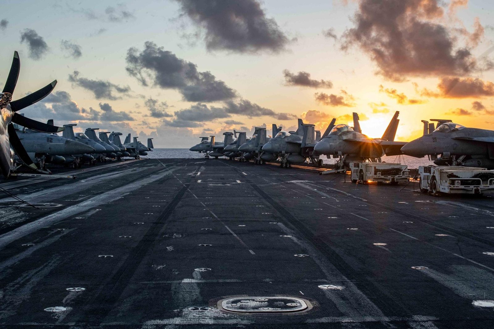 An aircraft carrier flight deck at sunset. Rows of parked fighter jets line both sides of an empty deck, and the low sun sits on the horizon.