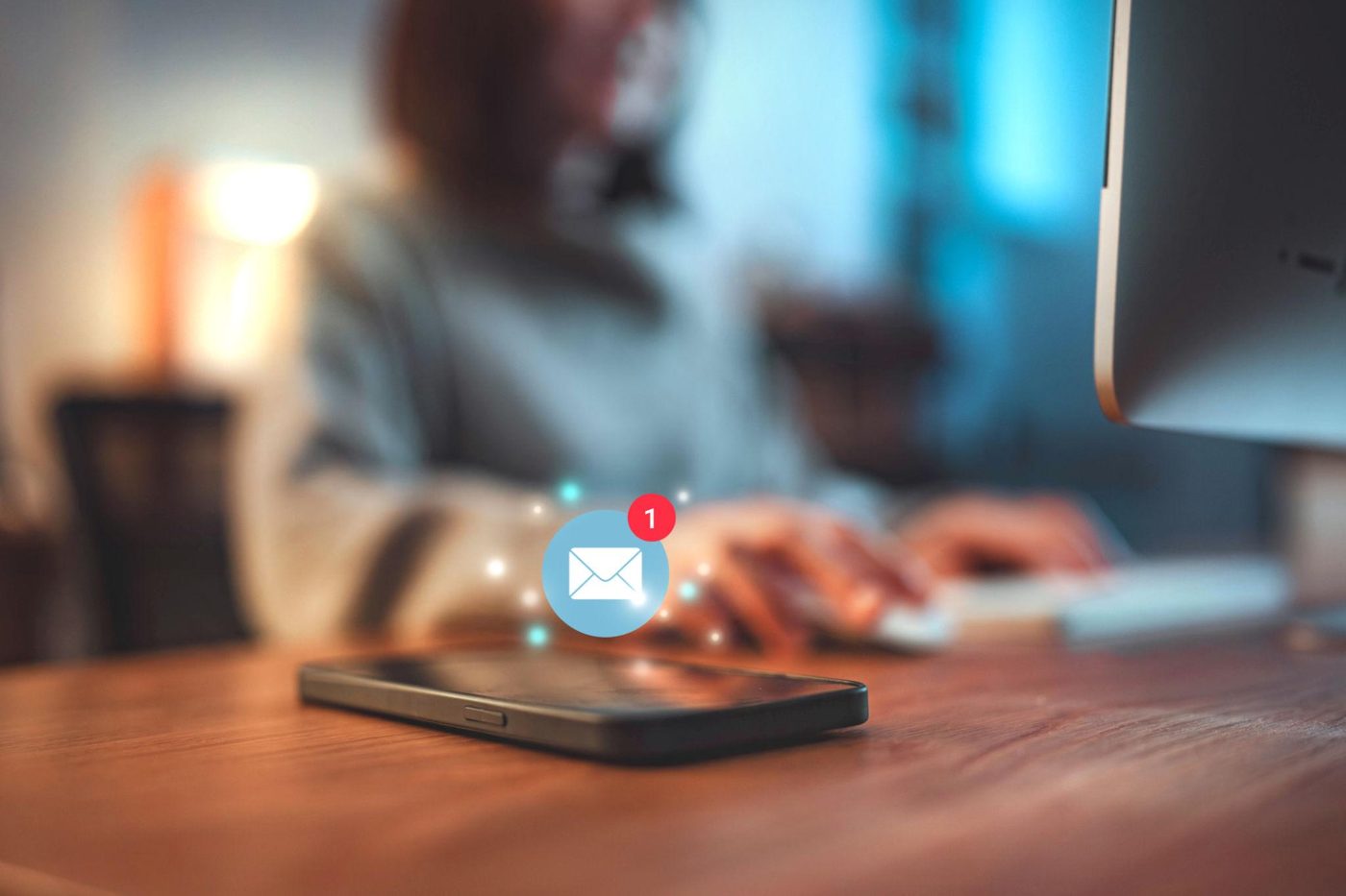 A smartphone lies on a wooden desk with a glowing mail icon and a red number 1 floating above it. Behind it, out of focus, a person works at a computer.