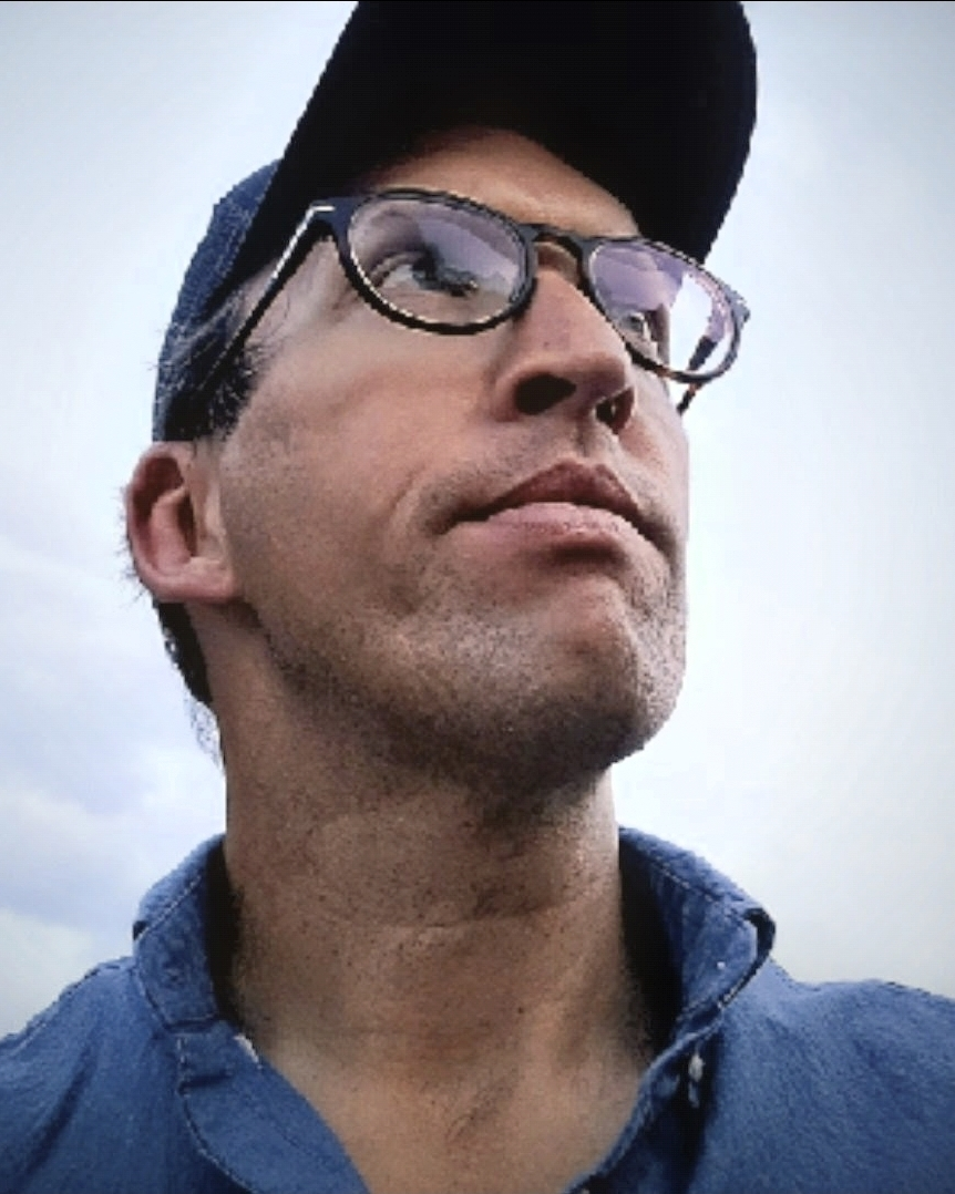 Mark Kantner, the practitioner — outdoors in a chambray shirt and cap, gaze lifted toward an open sky.